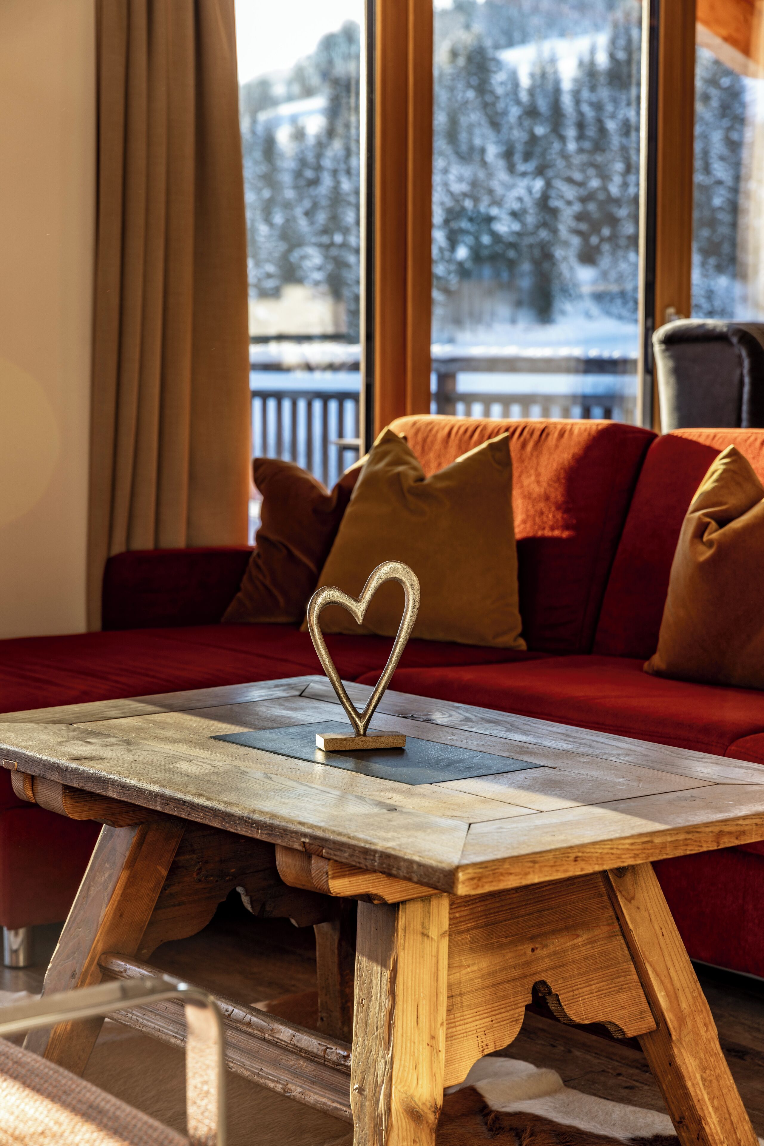 A cozy room with a wooden table, a red sofa, and a view of snowy trees through large windows.