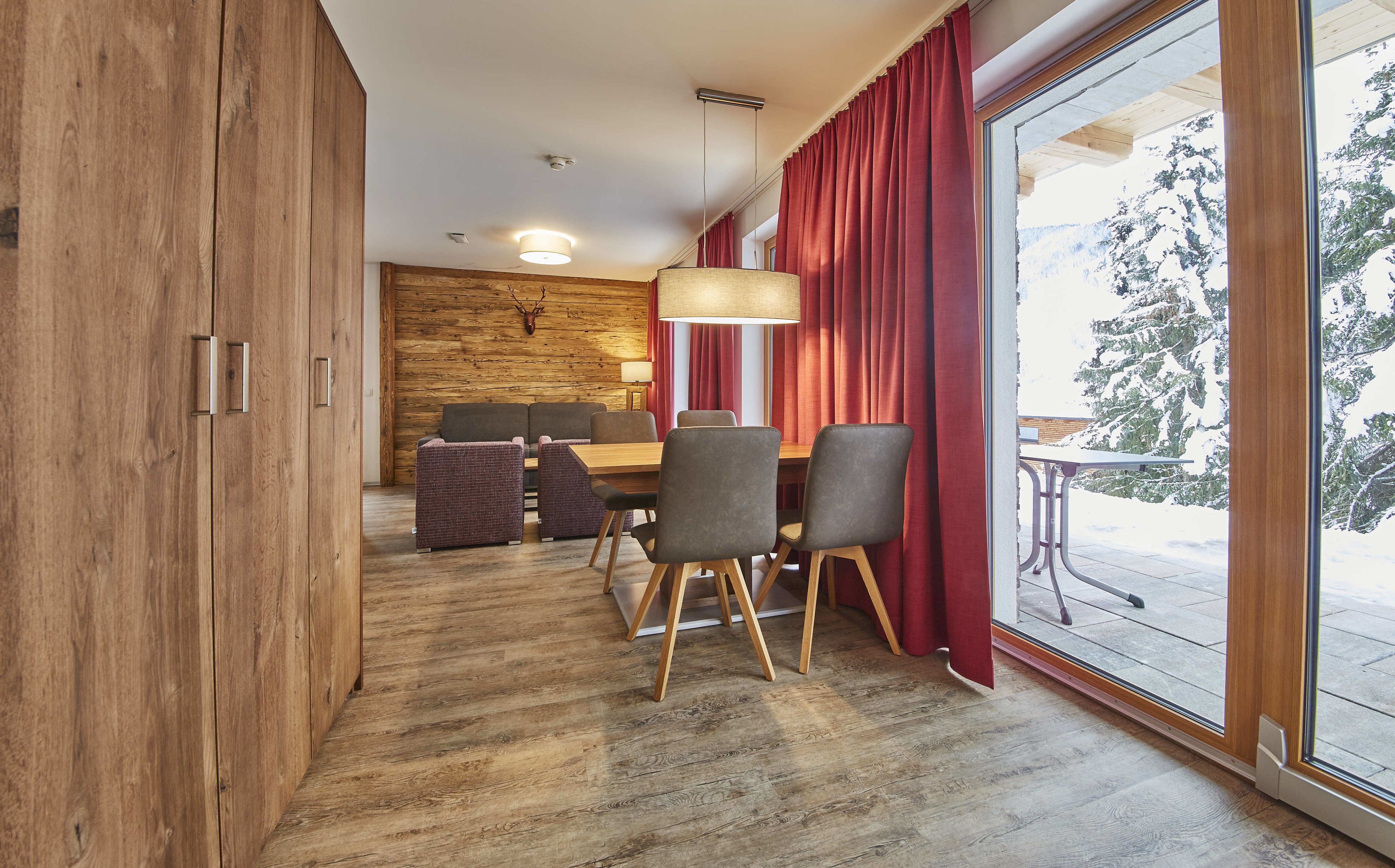 A dining area with a wooden table and chairs, a red curtain, and a view of a snowy landscape through a large window.