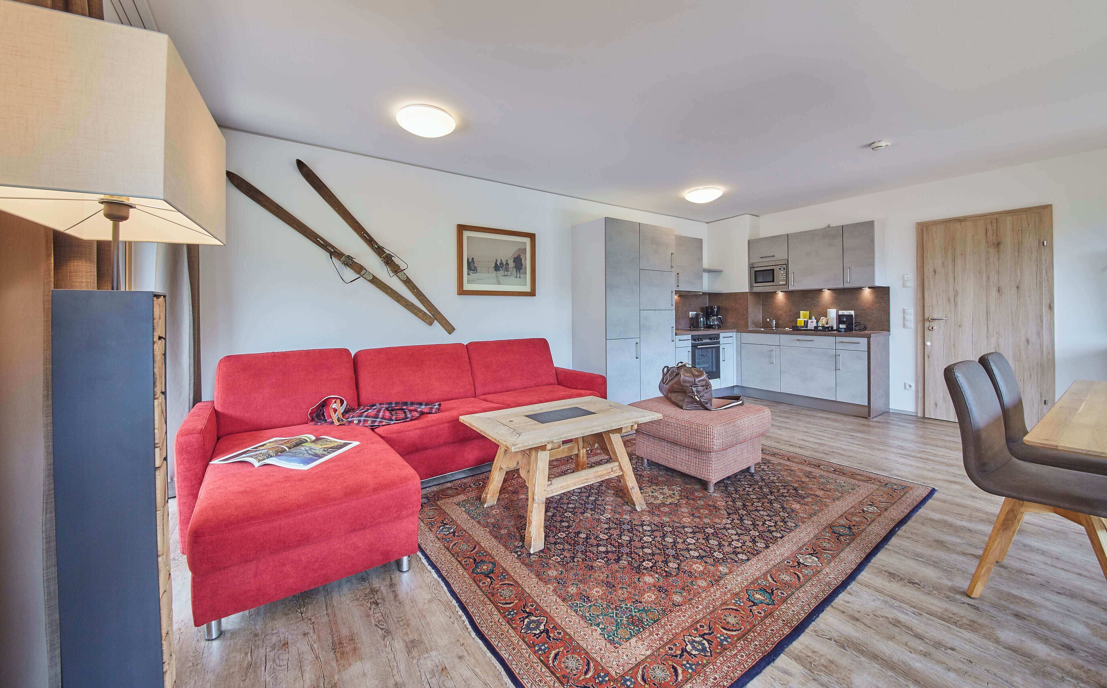A modern living room with a red sofa, a wooden coffee table, and a patterned rug. A kitchen with grey cabinets and a wooden door is visible in the background.
