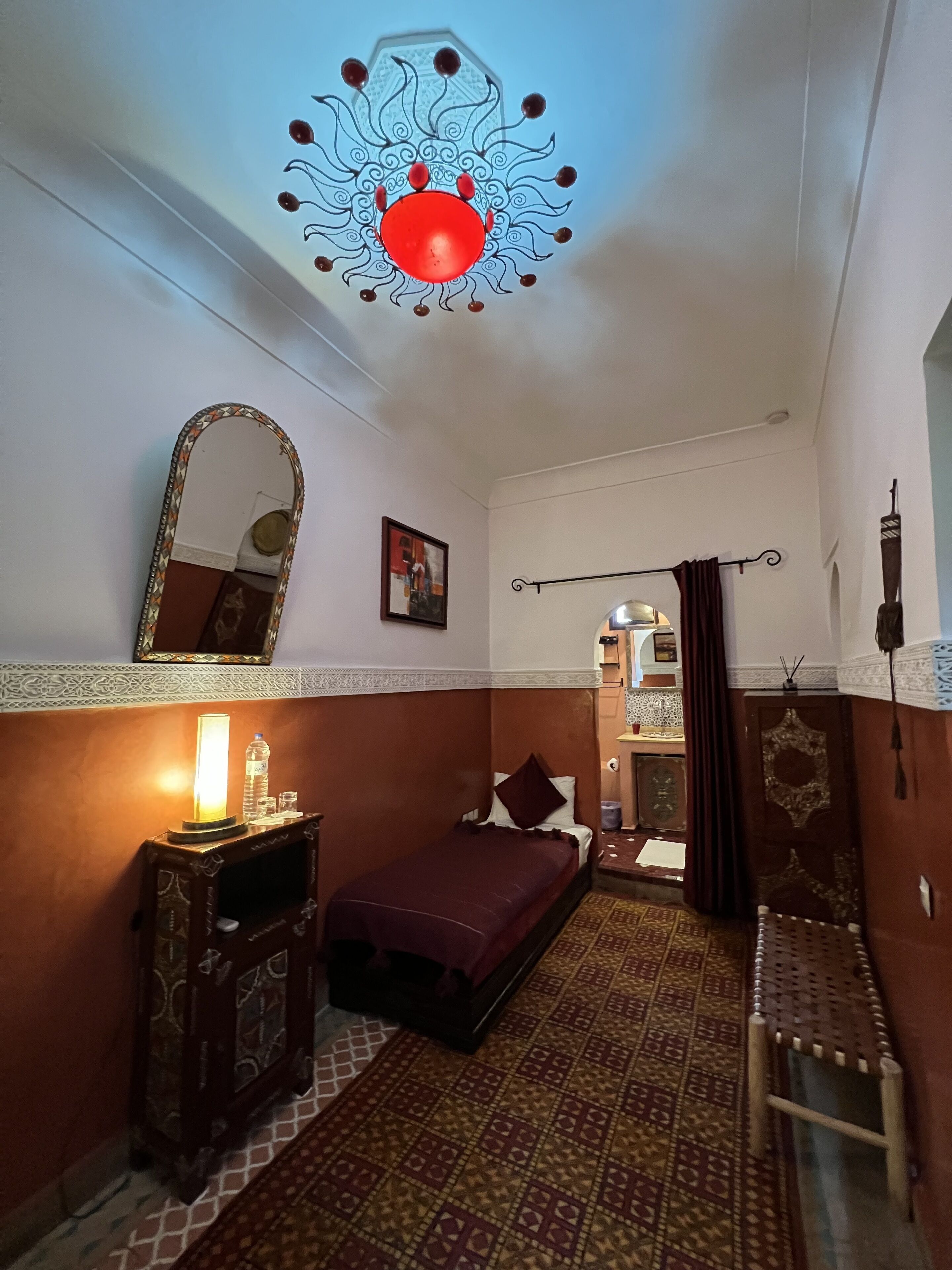 A small, cozy room with a bed, a bedside table with a lamp, a mirror, and a patterned rug.