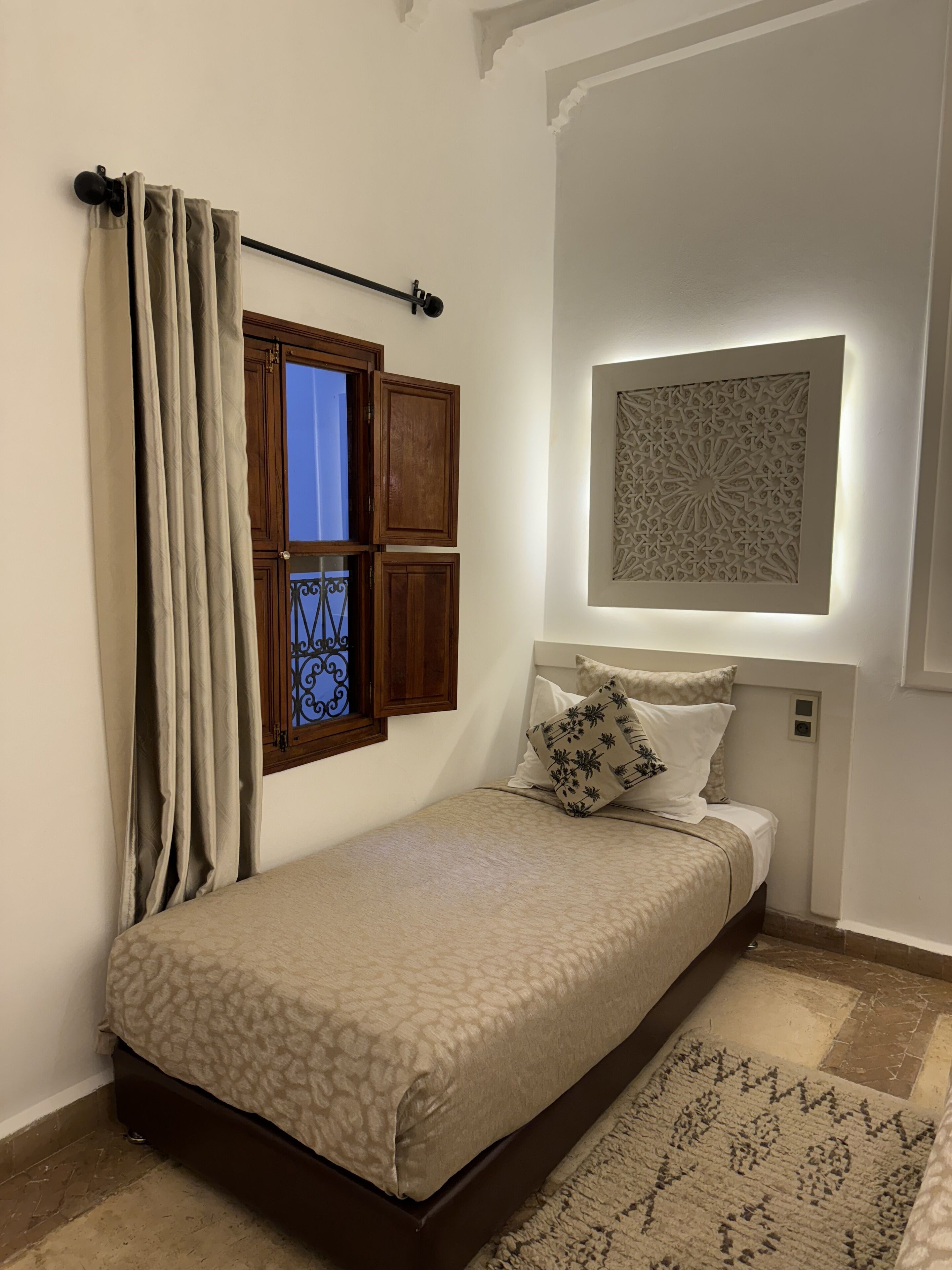 Quarto com cama, cortinas, janela com persianas, uma obra de arte na parede e tapete estampado.