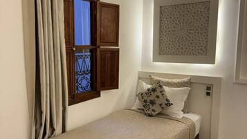 Quarto com cama, cortinas, janela com persianas, uma obra de arte na parede e tapete estampado.