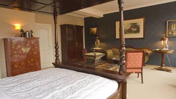 A four-poster bed with a white and gray patterned bedspread, a wooden wardrobe, a small table with a lamp, a chair, and a framed picture on the wall.