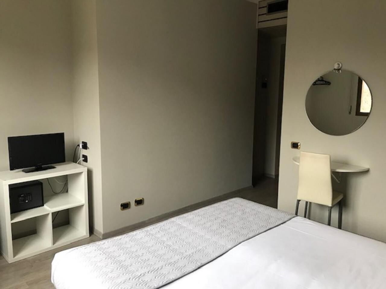 A hotel room with a bed, a TV on a stand, a small table, a chair, and a round mirror on the wall.