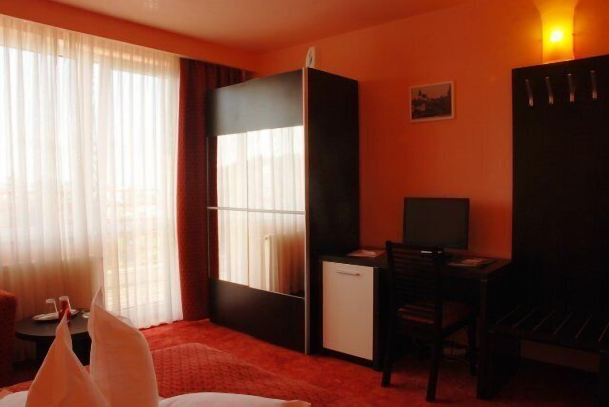 Quarto de hotel com uma cama, uma mesa, uma cadeira, um pequeno frigorífico e uma janela com cortinas.