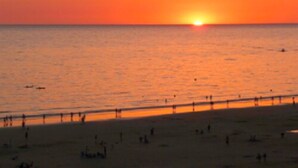Escena de playa con un atardecer sobre el océano, gente en la arena y un muelle al fondo.