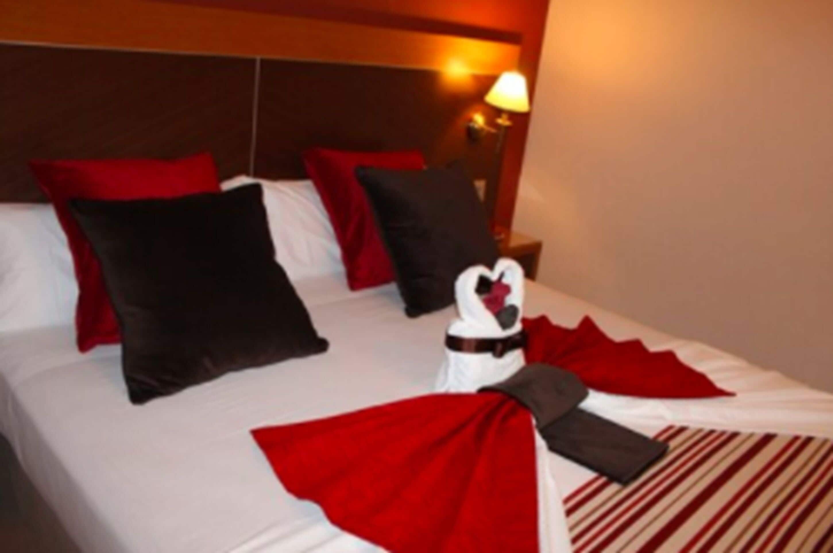 Quarto de hotel com cama bem arrumada, travesseiros vermelhos e pretos, toalha branca e colcha listrada em vermelho e branco.