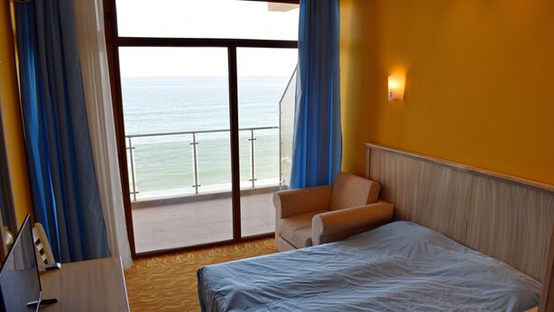 A hotel room with a bed, a chair, a desk with a computer, and a view of the ocean through a large window. - Brigantina Beach (Varna)