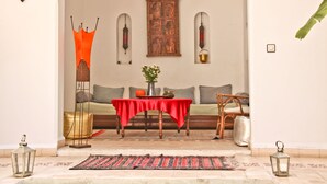 Una habitación con un sofá, una mesa con mantel rojo, un adorno de pared y dos faroles fijados a la pared.