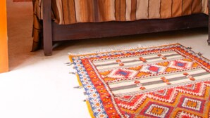 Una cama con dosel, un edredón a rayas, una alfombra colorida y una mesita de noche con un jarrón de flores.