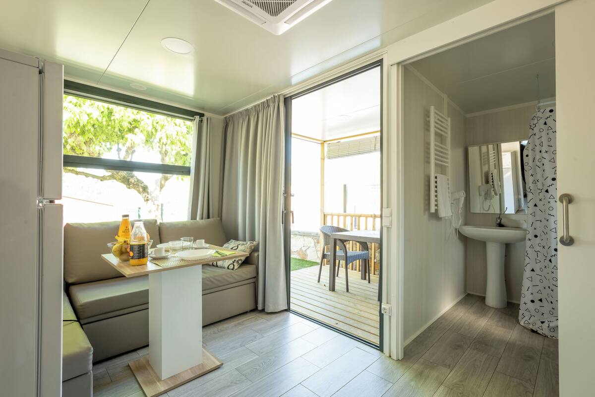Quarto moderno com sofá, uma pequena mesa com uma garrafa, uma cadeira e vista para um deck externo.