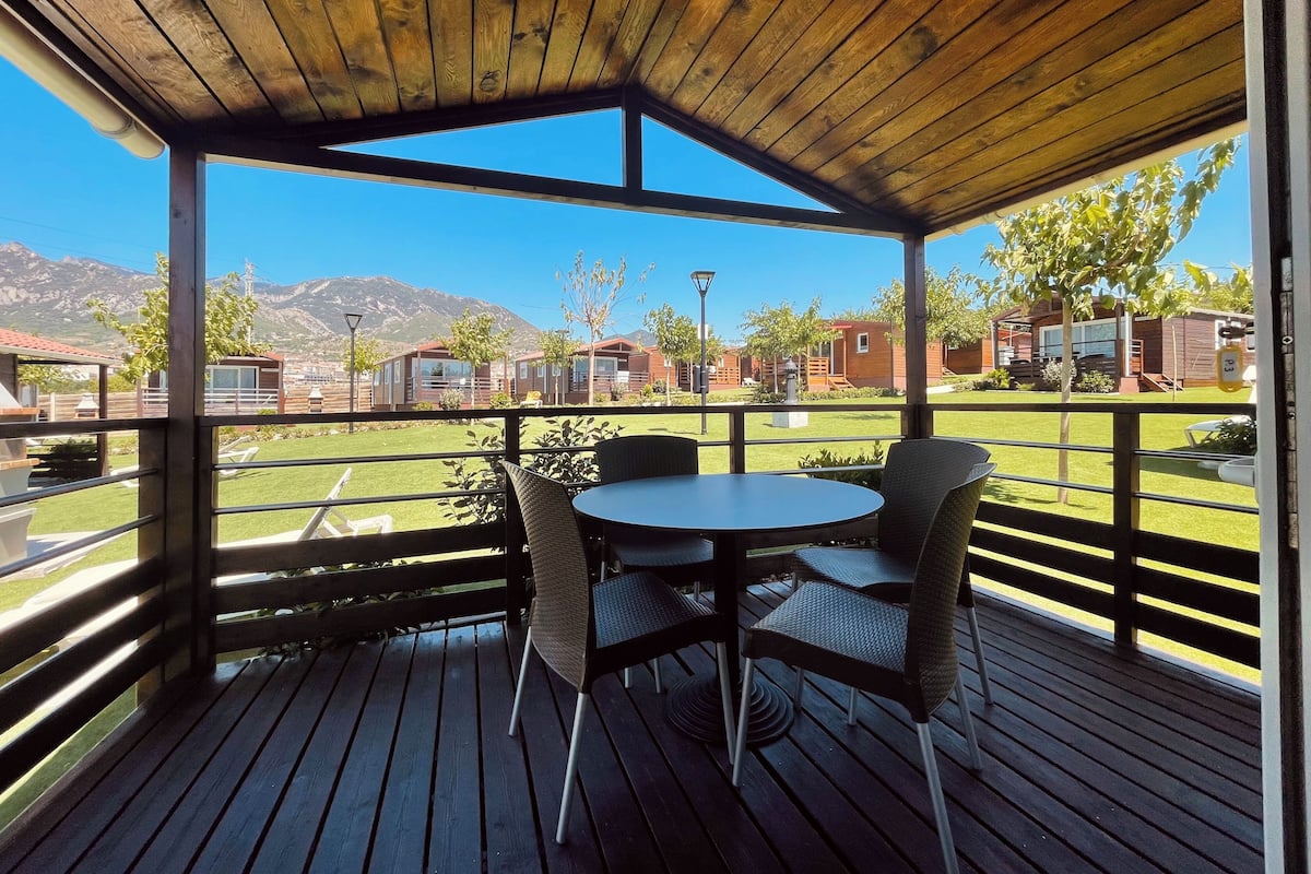 Uma área de estar coberta ao ar livre, com mesa e cadeiras, com vista para um gramado bem cuidado e uma fileira de cabanas.