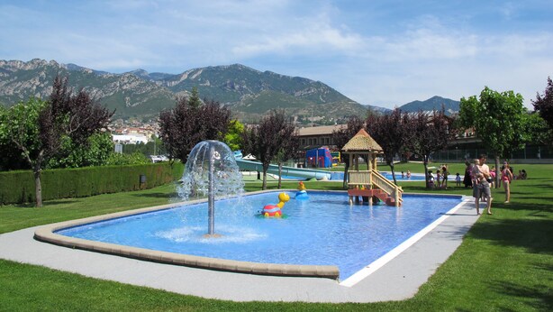 Una piscina con una fuente de agua, rodeada de césped y árboles, con montañas al fondo.