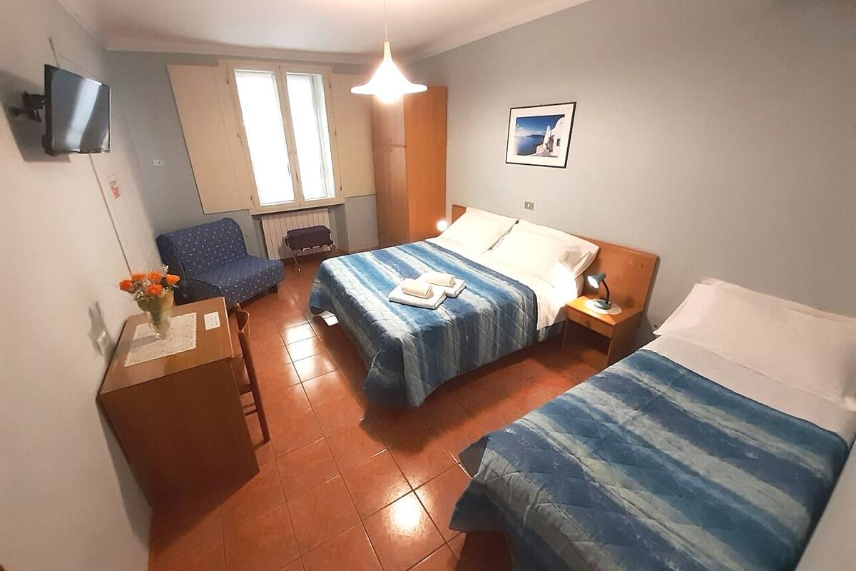 Una habitación pequeña y bien iluminada con dos camas, un escritorio con silla, un televisor y una ventana.