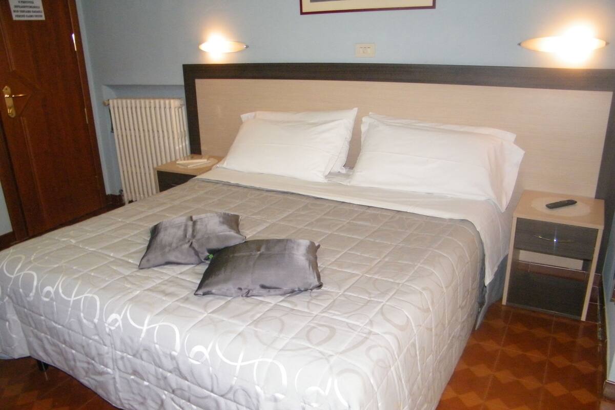 Una cama bien hecha con una colcha acolchada y dos almohadas, una mesita de noche con un control remoto y una lámpara de pared.