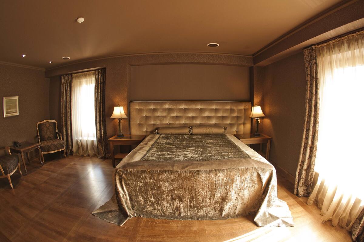 A spacious bedroom with a large bed, two bedside tables, a chair, and a window with curtains.