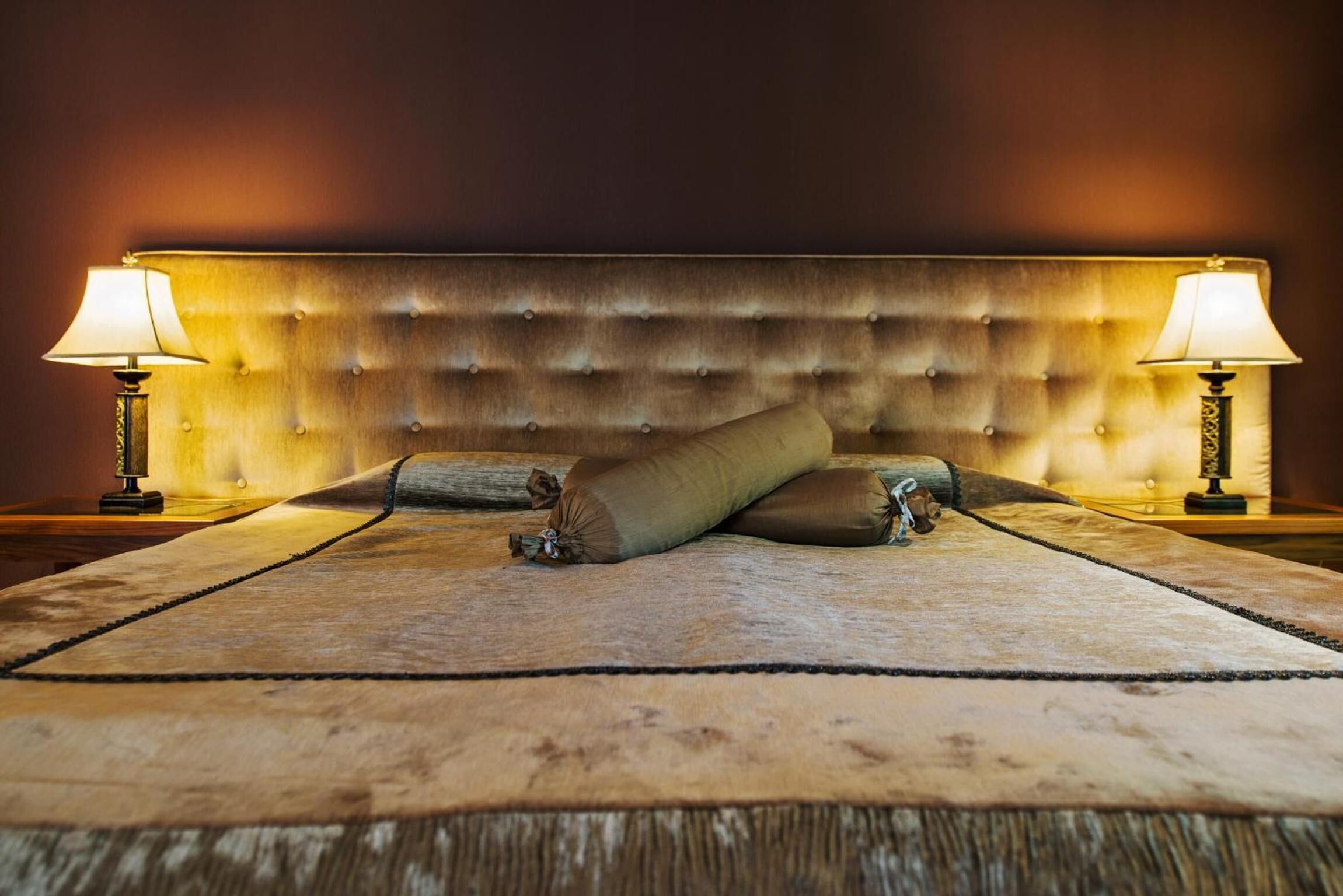 A bed with a tufted headboard, two bedside lamps, and a rolled-up blanket on a textured rug.