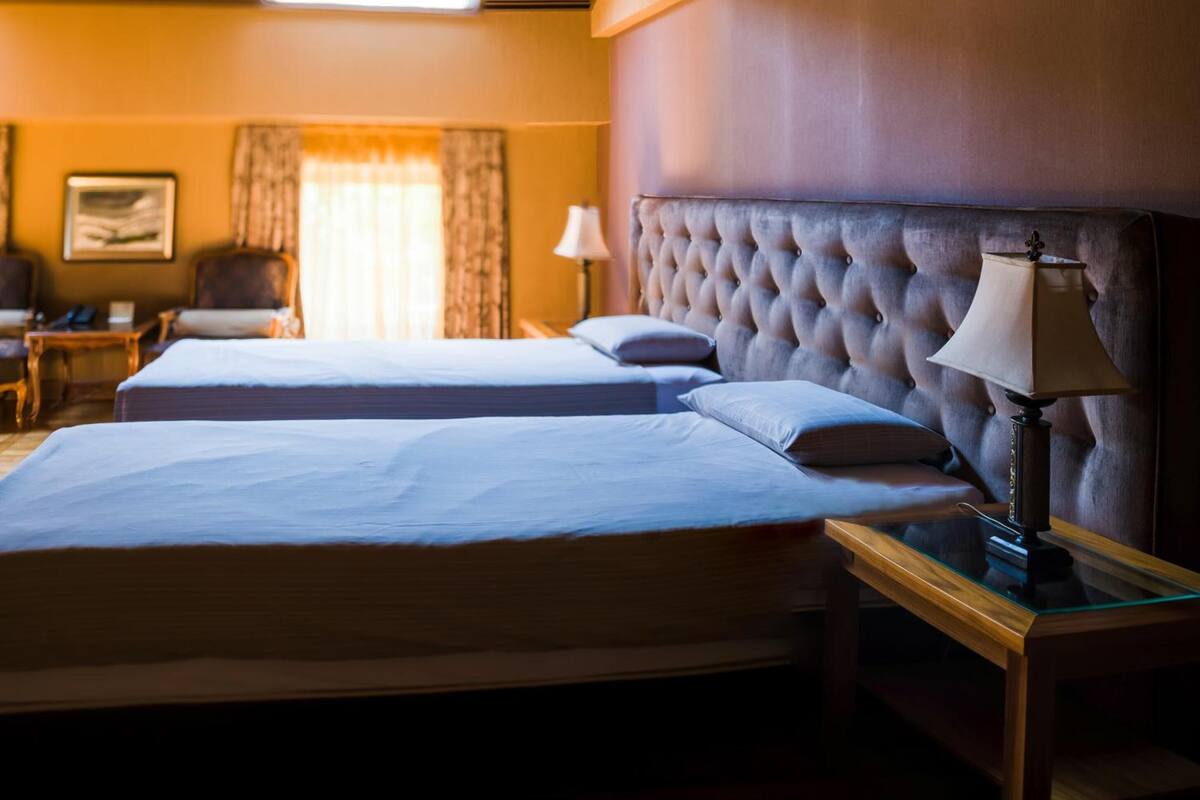 A hotel room with two beds, a large tufted headboard, a bedside lamp, a chair, and a desk.