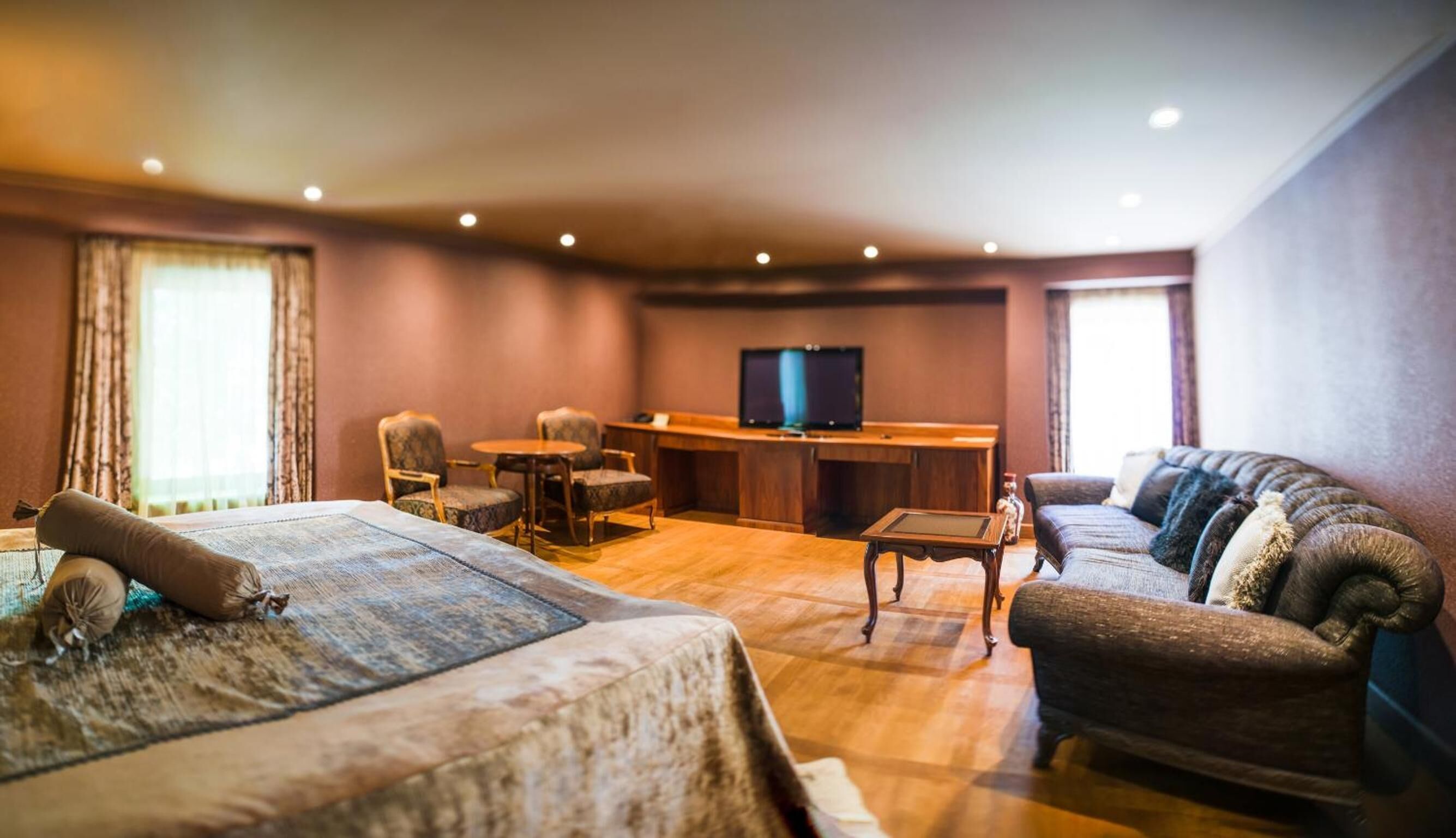 A hotel room with a bed, a sofa, a desk with a TV, and a dining table.