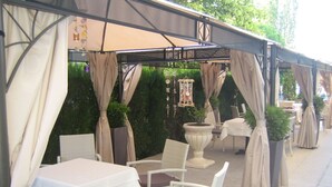 Un patio al aire libre con mesas y sillas, cubierto por una pérgola con cortinas.