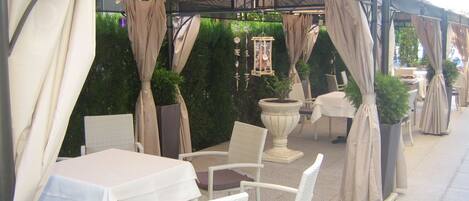 Un espace extérieur aménagé en terrasse, avec des tables et des chaises, recouvert d’un auvent agrémenté de rideaux.