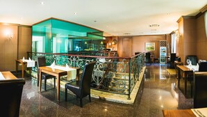 Vestíbulo de hotel con una escalera central, un comedor con mesas y sillas, y vista de la cocina a través de un panel de vidrio.