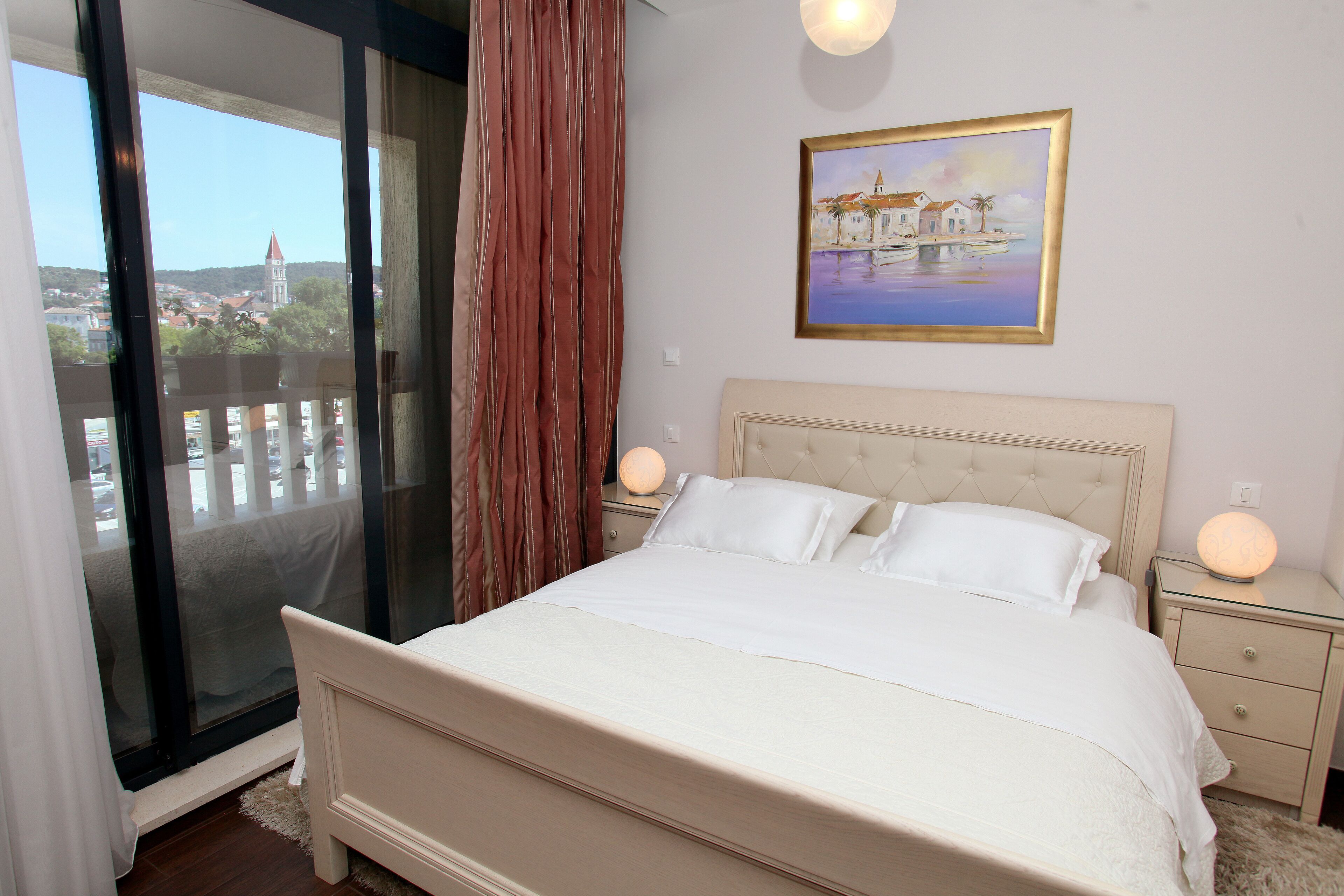Ein Schlafzimmer mit einem Bett, Nachttischen, einem Gemälde und Blick auf eine Stadt durch ein Fenster.