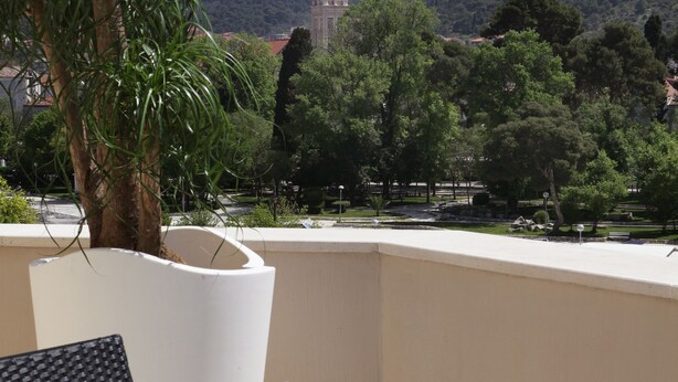 Un balcón con una mesa dispuesta para dos personas, mostrando un plato de fruta y dos vasos de una bebida amarilla. Se ve una palmera y un edificio con techo rojo al fondo.