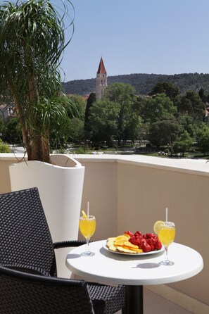 A balcony with a table set for two, featuring a plate of fruit and two glasses of a yellow beverage. A palm tree and a building with a red roof are visible in the background. - Hotel Bellevue (Trogir)