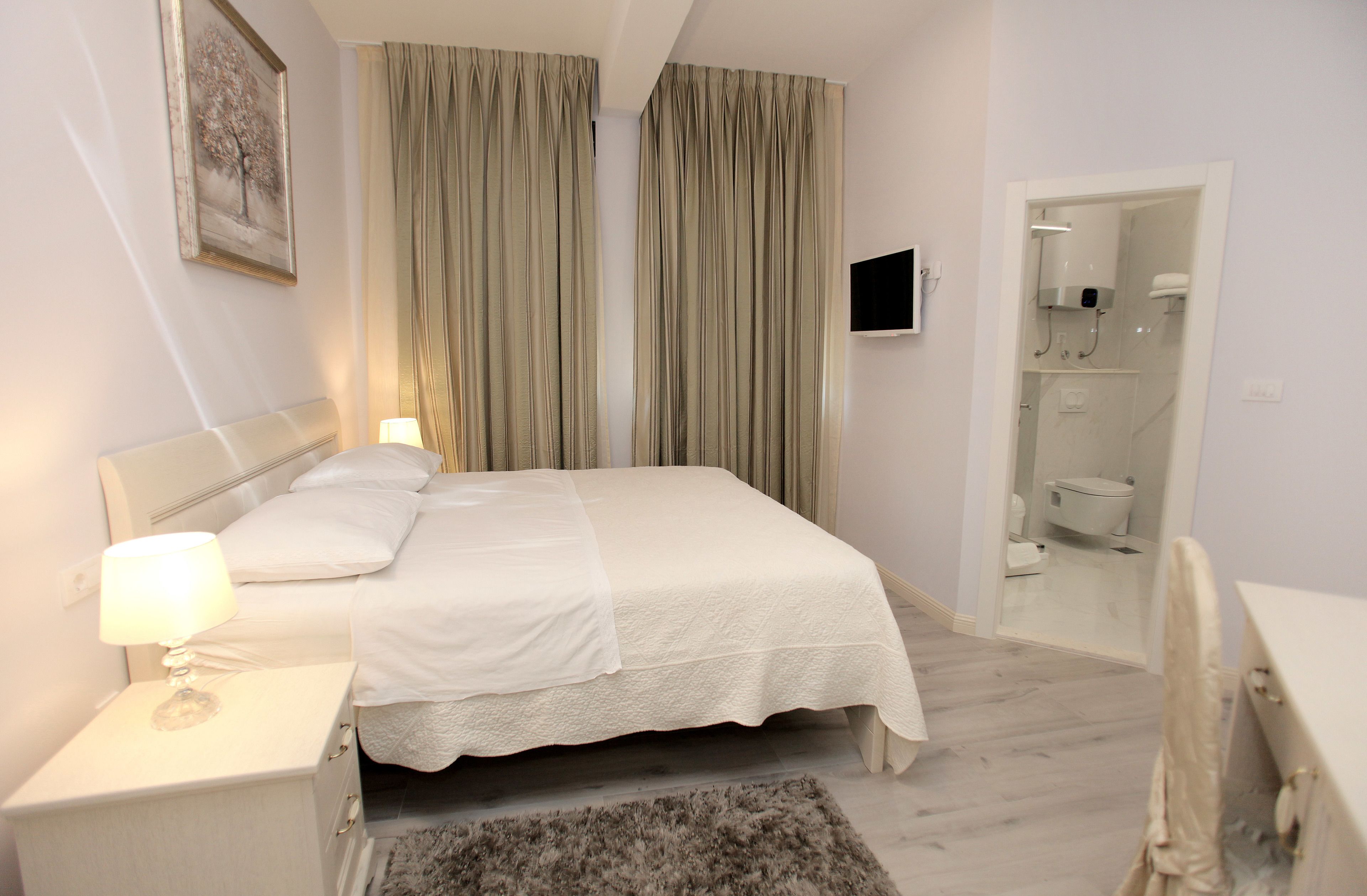 A hotel room with a bed, bedside table, lamp, curtains, a television, and a bathroom visible through an open door.