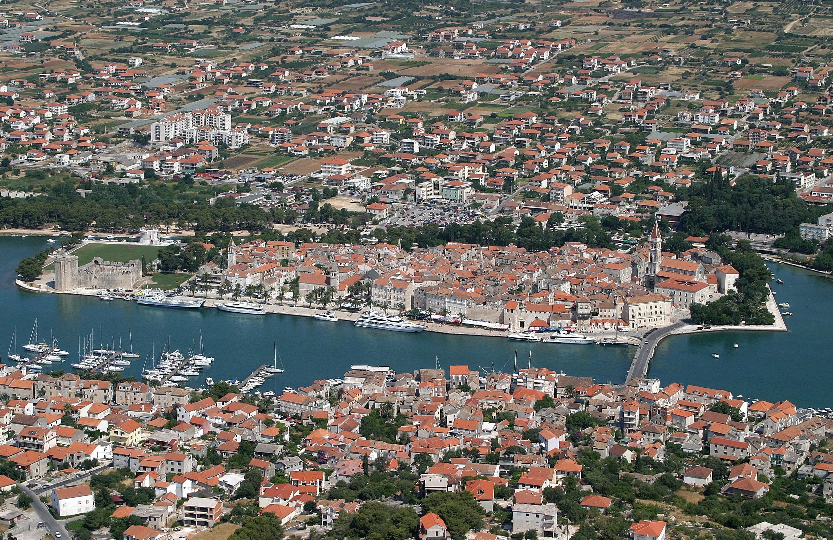 Vue aérienne d’une ville côtière, avec un ensemble dense de bâtiments aux toits rouges, une église imposante et un port de plaisance rempli de bateaux.
