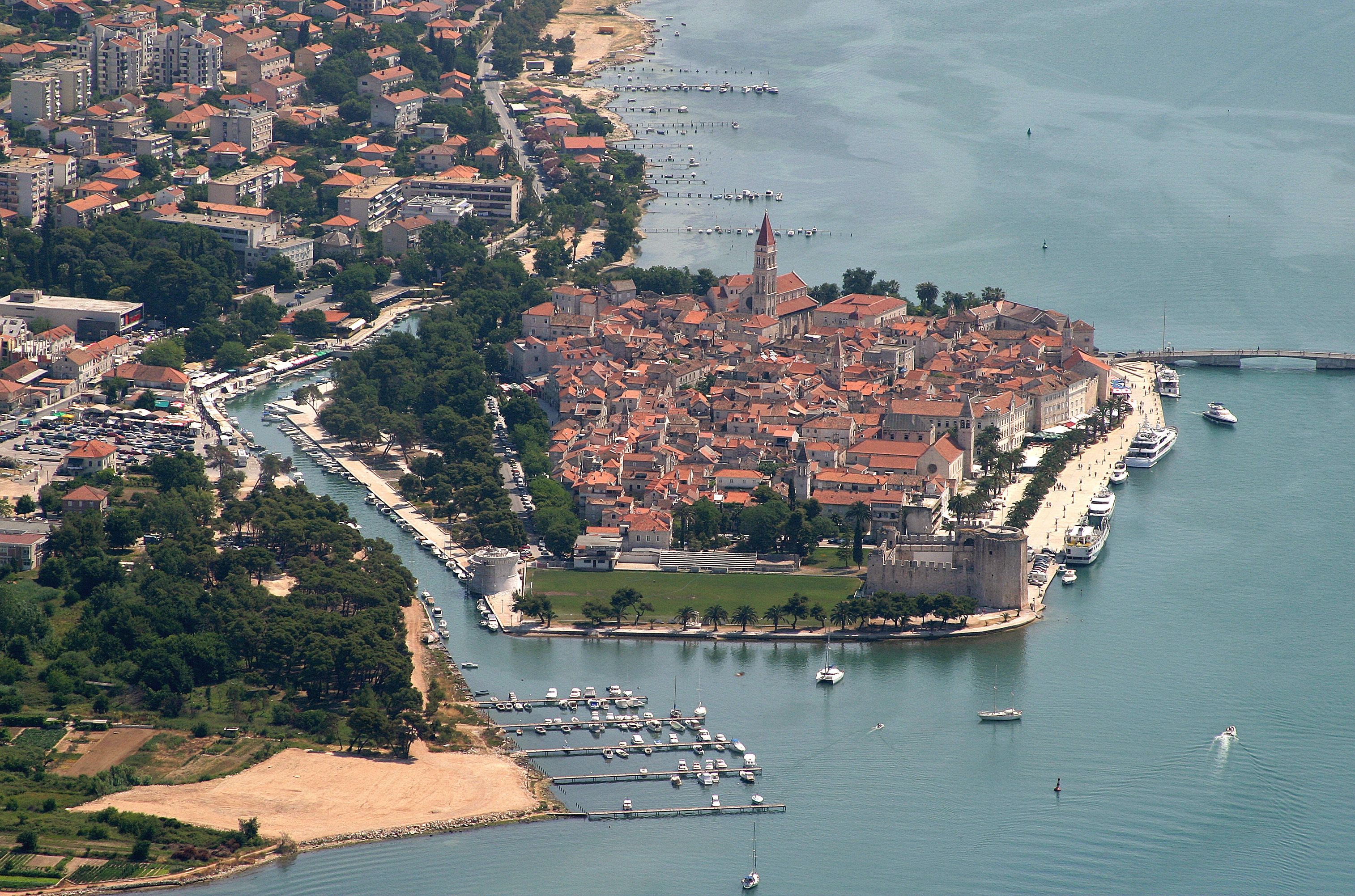 Vista aérea de un pueblo costero con una torre destacada, un puerto deportivo con barcos, y una mezcla de edificios residenciales y comerciales.