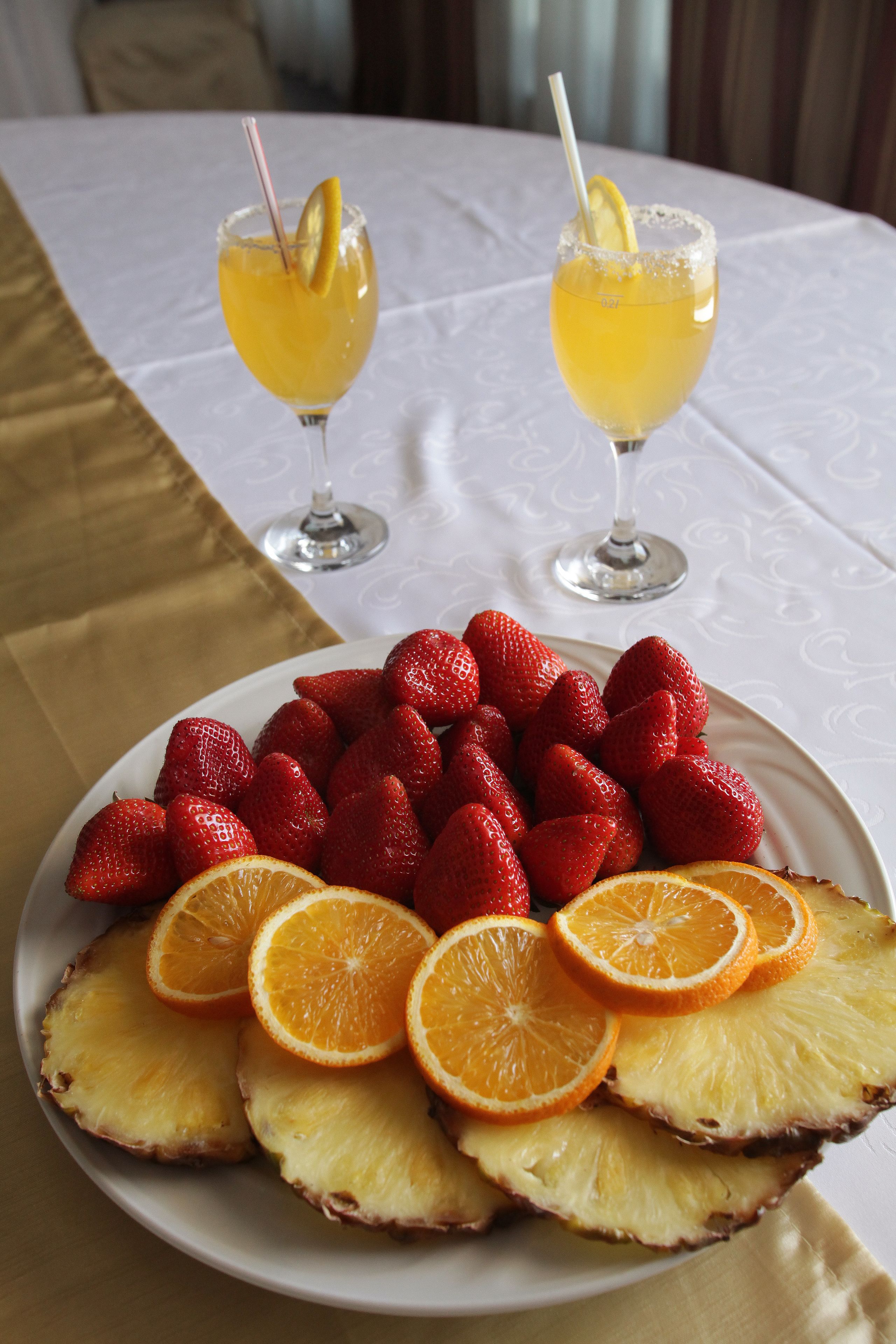A table with a white tablecloth, a plate of strawberries, orange slices, and pineapple slices, and two glasses of a yellow beverage with a straw and a lemon slice.