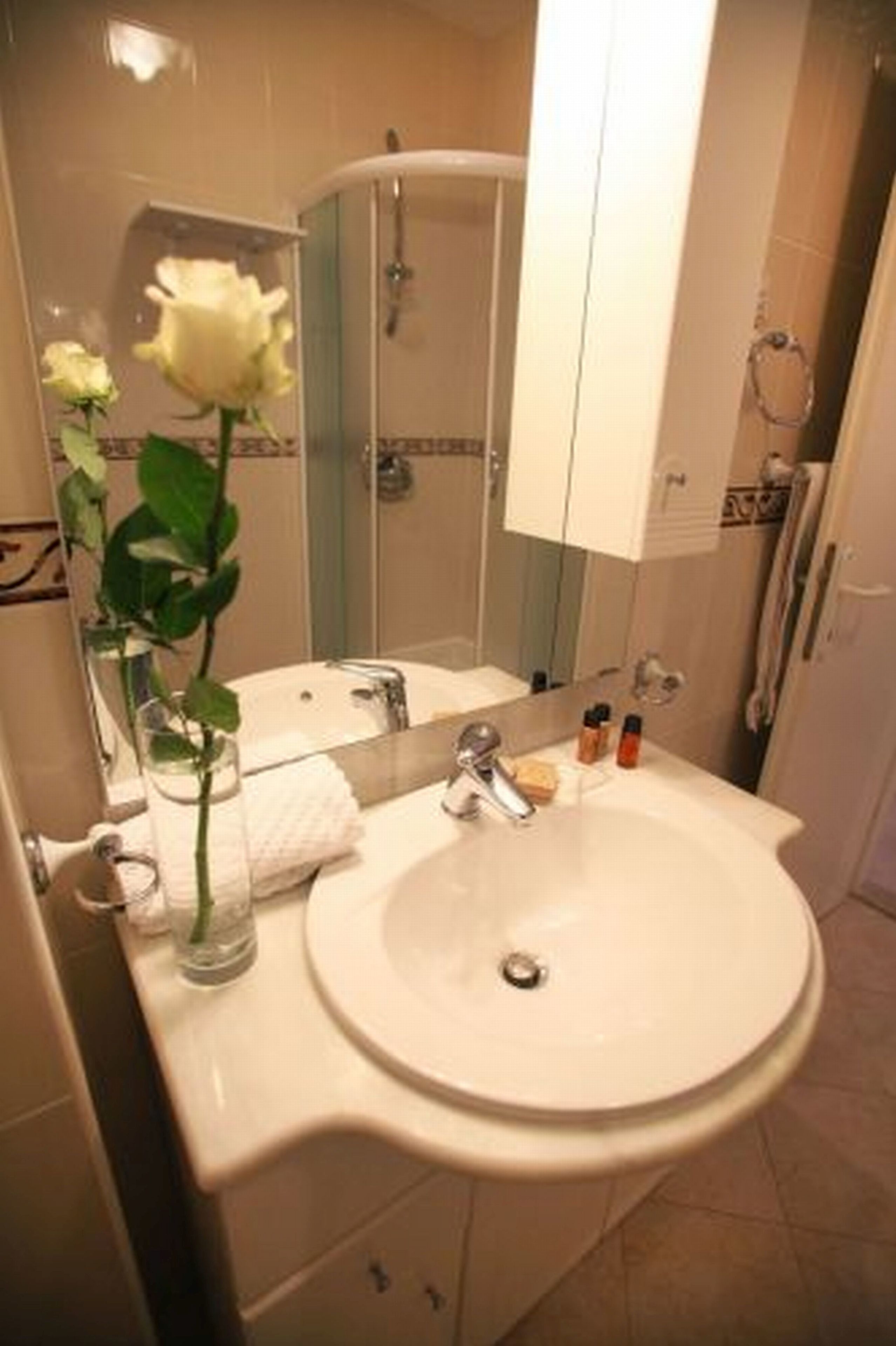 Une salle de bain avec un lavabo blanc, un miroir, un vase contenant une rose blanche et des articles de toilette.