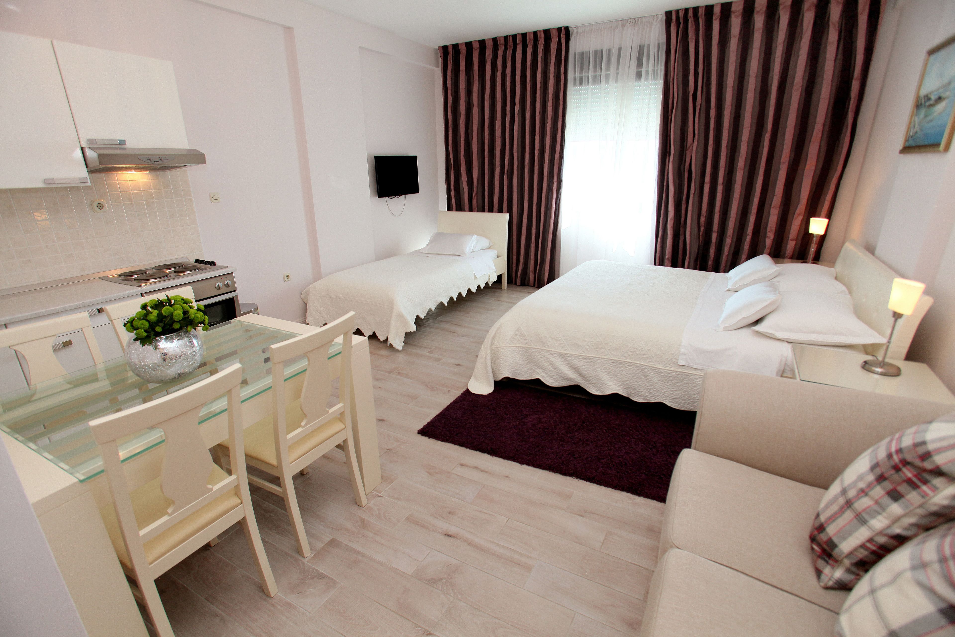 Une chambre d’hôtel avec une kitchenette, un lit avec du linge de lit blanc, une petite table avec des chaises et un téléviseur fixé au mur.