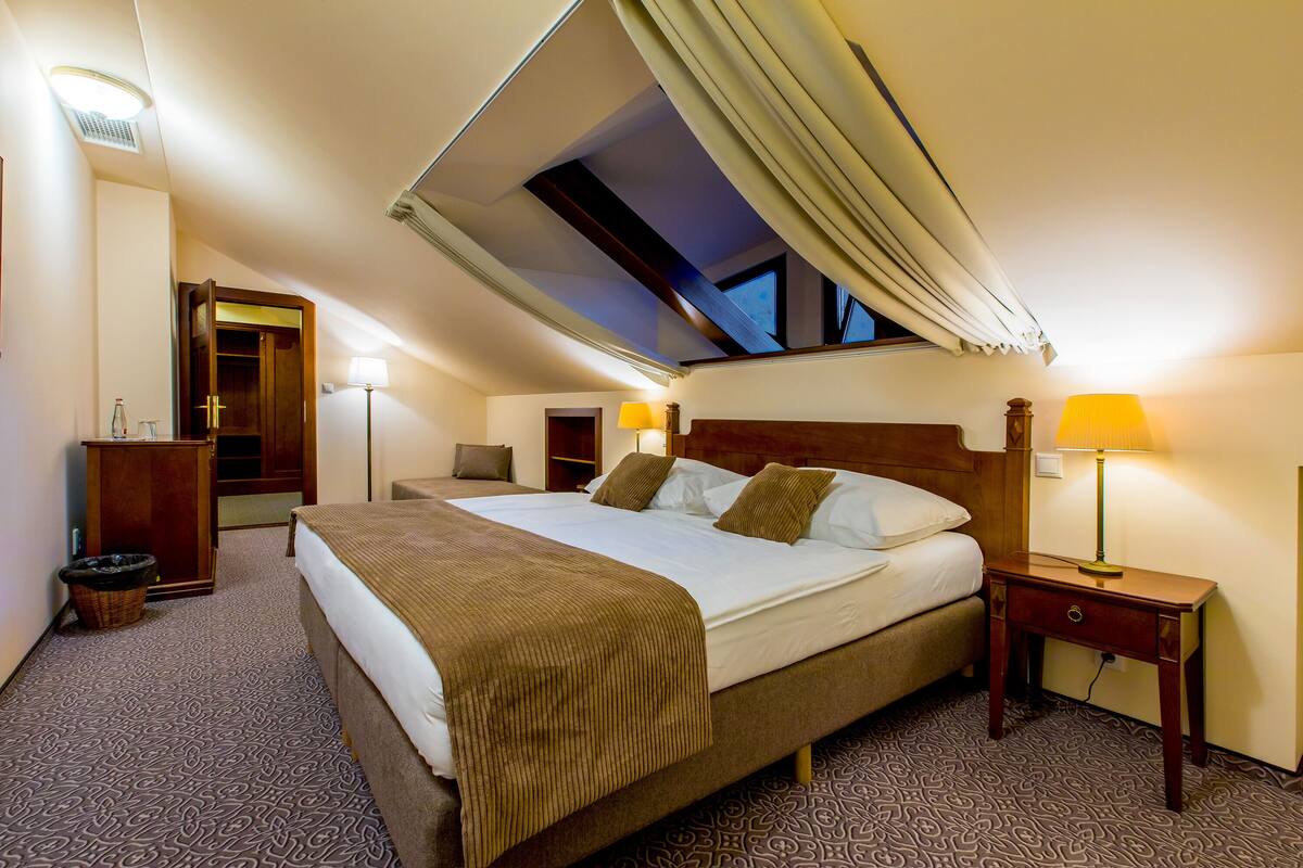 Habitación de hotel con una cama grande, dos mesitas de noche con lámparas, un armario de madera, un pequeño cubo de basura y un tragaluz en el techo.