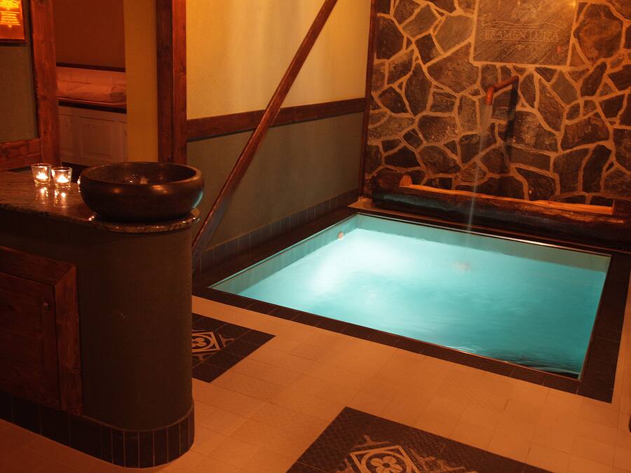 Un espacio de spa con pared de piedra, un banco de madera, una pequeña mesa con un cuenco y piso de baldosas.