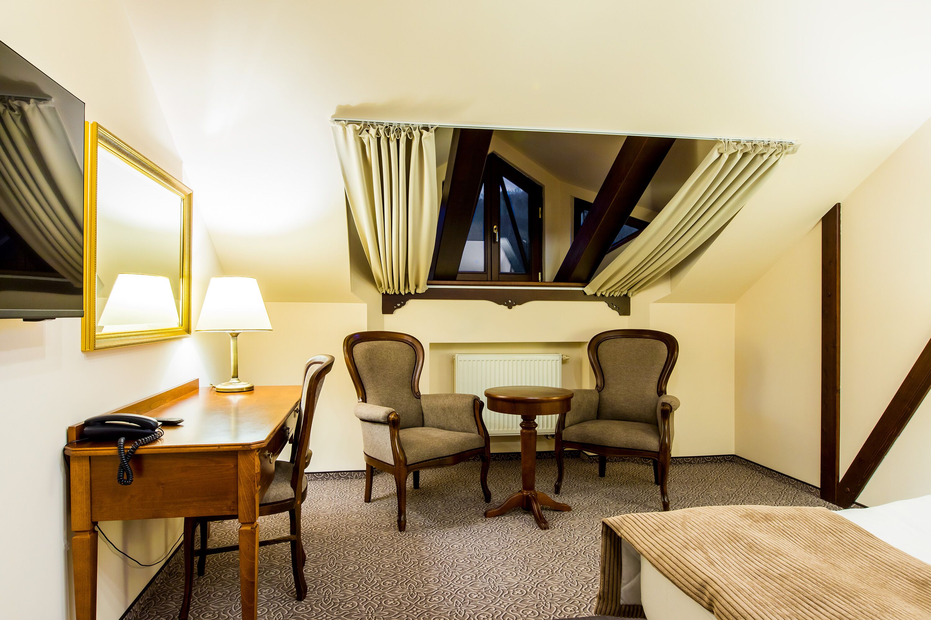 A hotel room with a desk, two armchairs, a small table, a mirror, and a window with curtains.