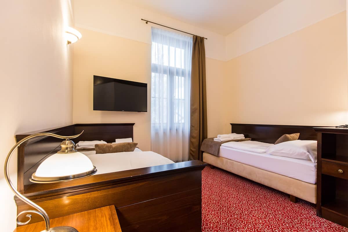 Una habitación de hotel con un escritorio de madera, una cama, otra cama adicional, un televisor montado en la pared y un ventanal con cortinas.