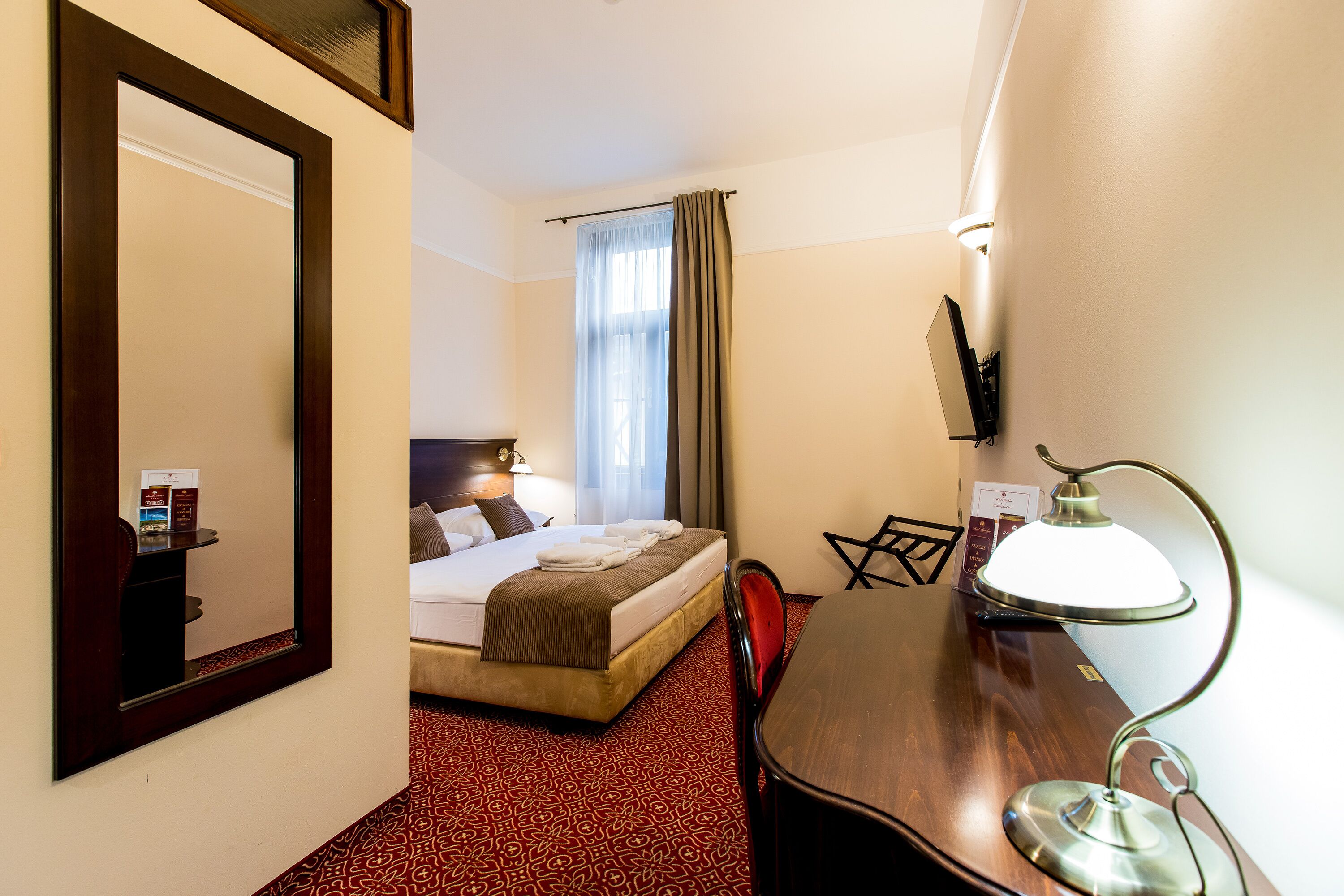 Hotelkamer met een bed, bureau met lamp, stoel, grote spiegel, raam met gordijnen en een televisie aan de muur.