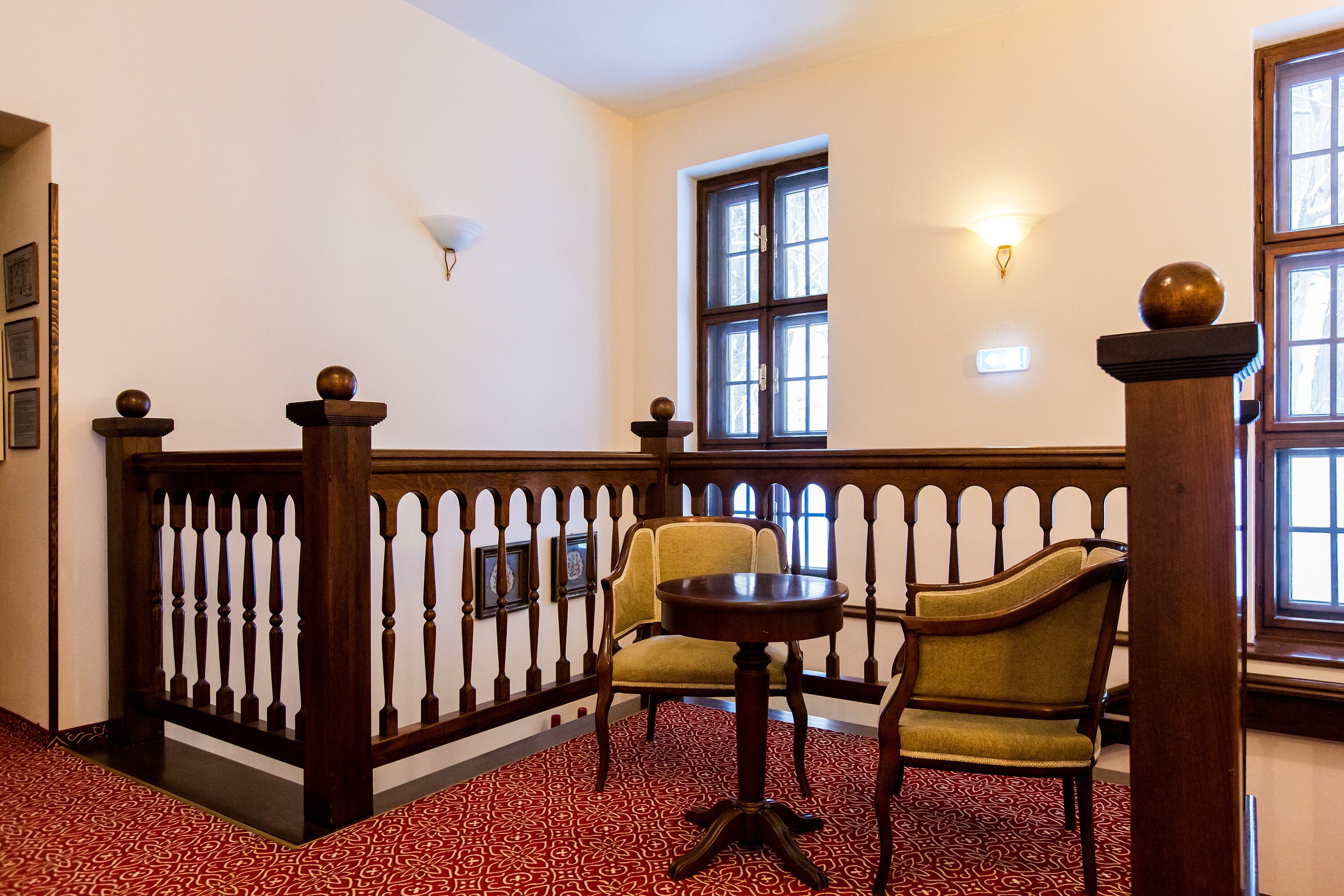 A hallway with a wooden railing, two chairs, a small round table, and a patterned carpet.