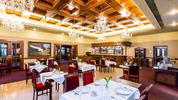 A restaurant with multiple tables, red chairs, chandeliers, and a bar area.