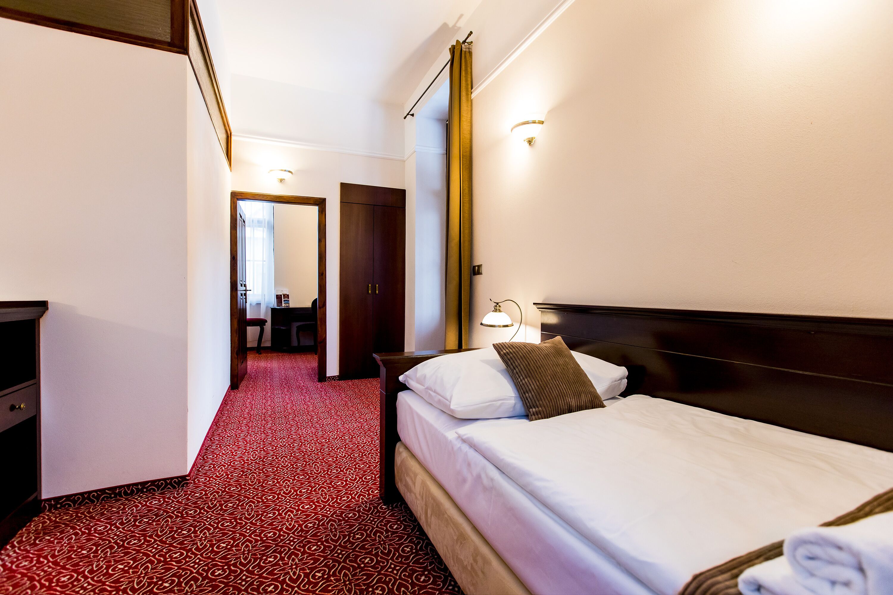 A hotel room with a neatly made bed, a desk, and a chair. The room has a red carpet and a wooden headboard.