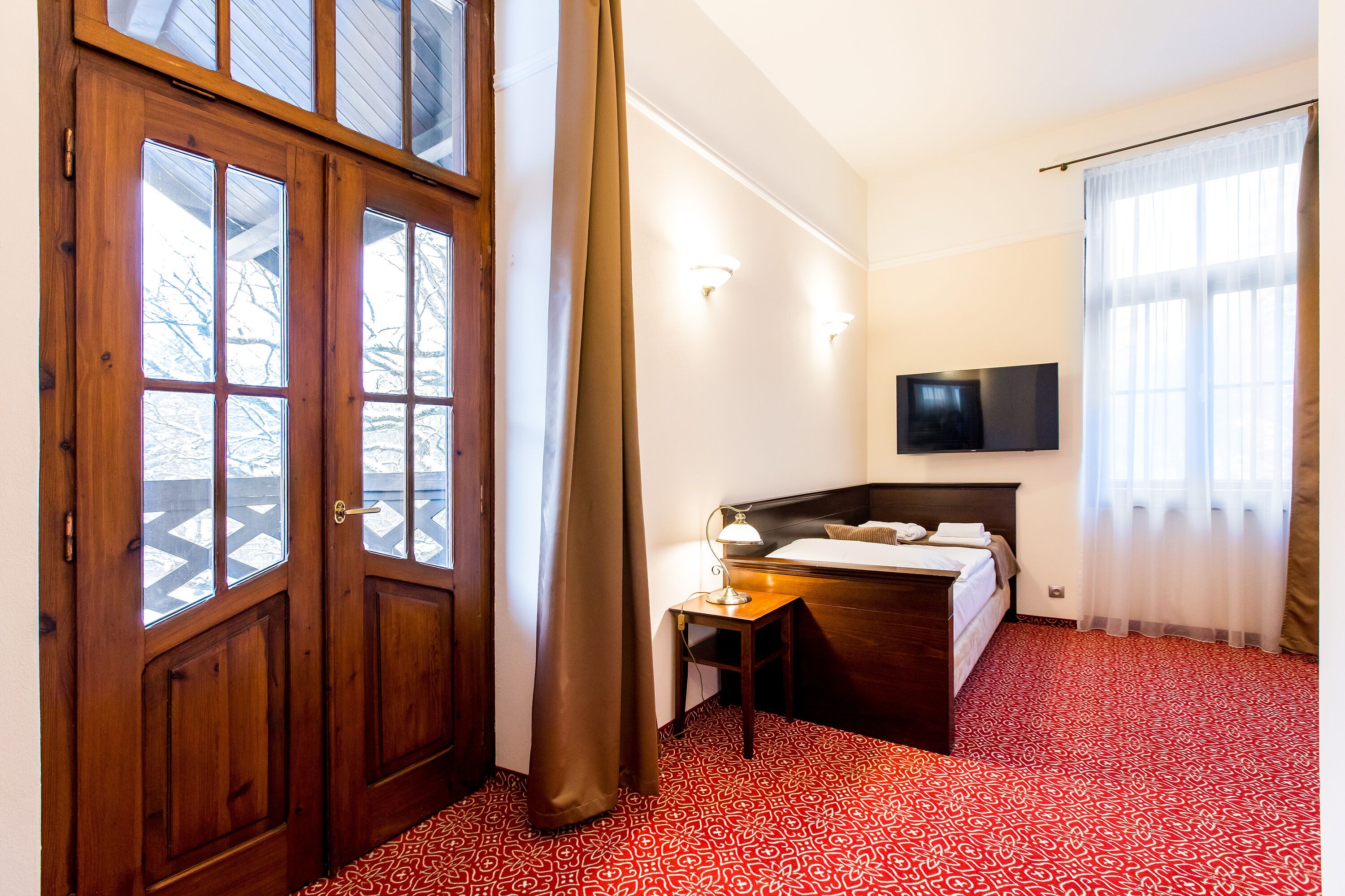 A hotel room with a wooden door, a bed with a headboard, a desk with a lamp, and a TV mounted on the wall.