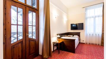A hotel room with a wooden door, a bed with a headboard, a desk with a lamp, and a TV mounted on the wall.