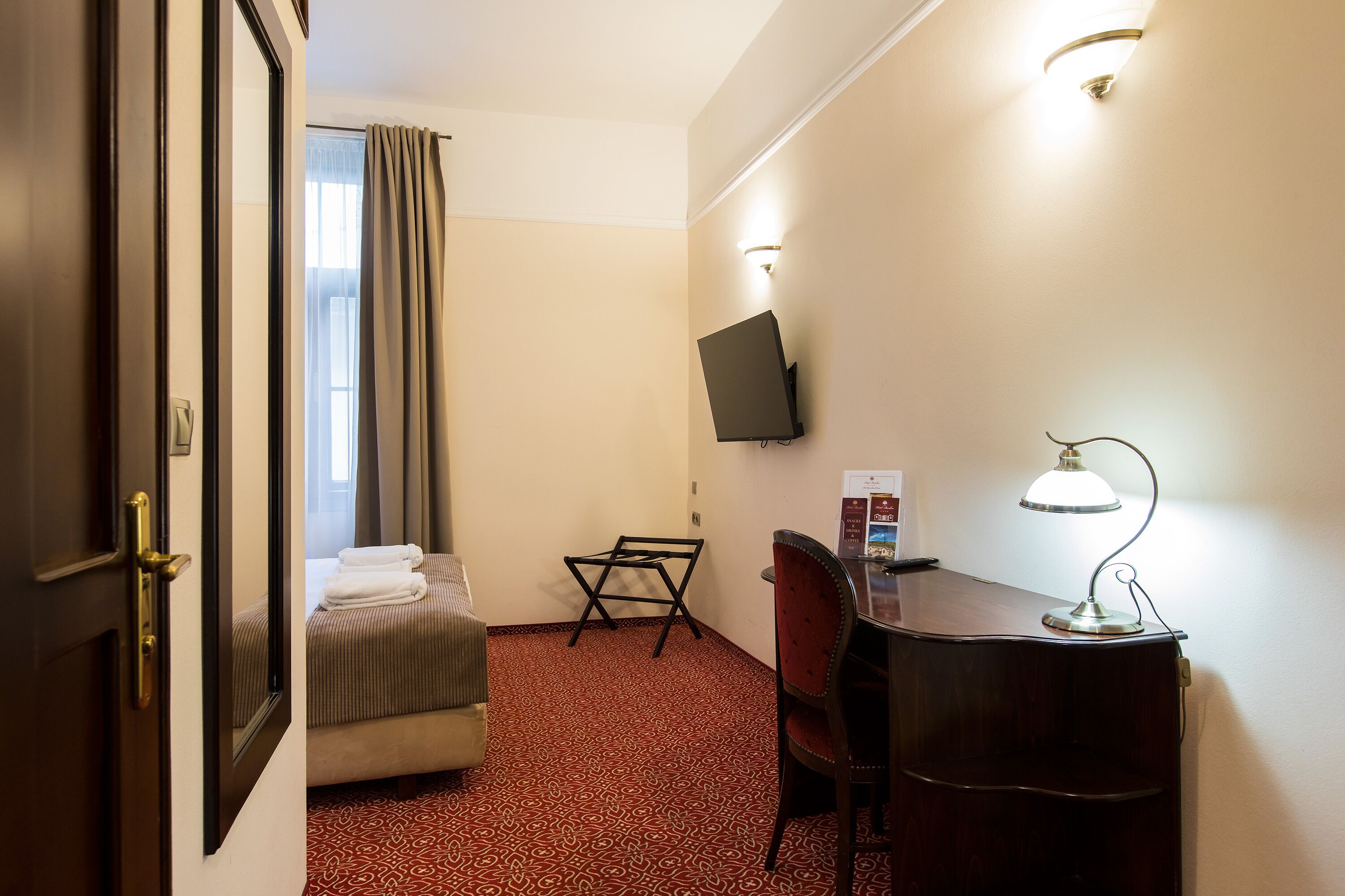 Habitación de hotel con una cama, un escritorio con lámpara, un televisor en la pared y una alfombra estampada.