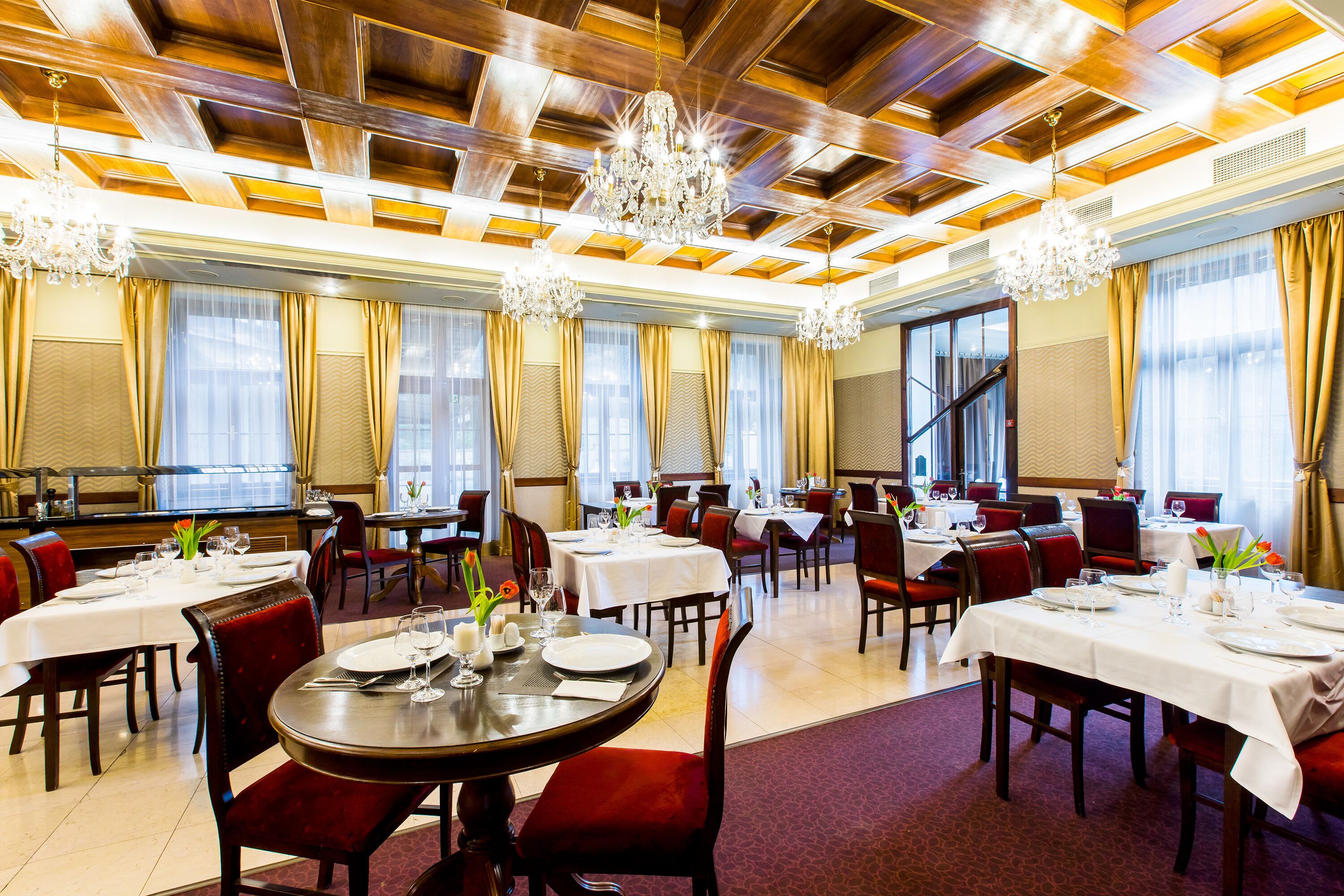 A spacious dining area with multiple tables set for dining, elegant chandeliers, and a high ceiling with wooden paneling.