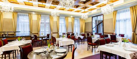 Un comedor amplio con varias mesas dispuestas para comer, candelabros elegantes y un techo alto con paneles de madera.