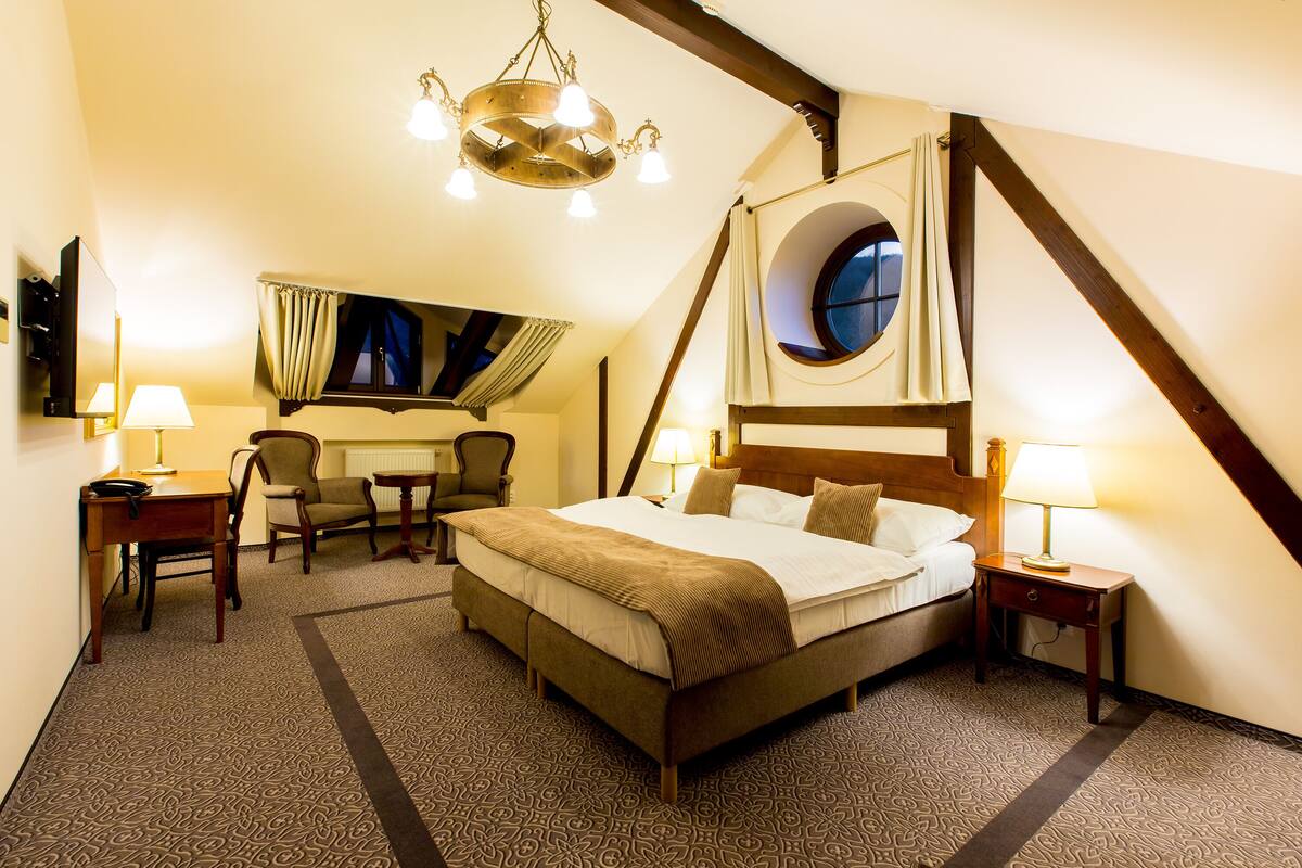 Habitación de hotel con una cama grande, un escritorio, dos sillas, una mesita y un ventanal circular, además de una araña.