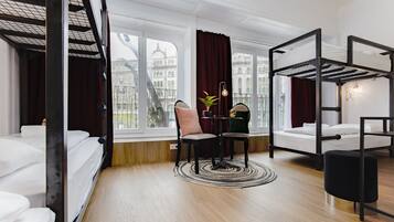 A hotel room with a bunk bed, a desk with a chair, a potted plant, and a view of buildings through large windows.